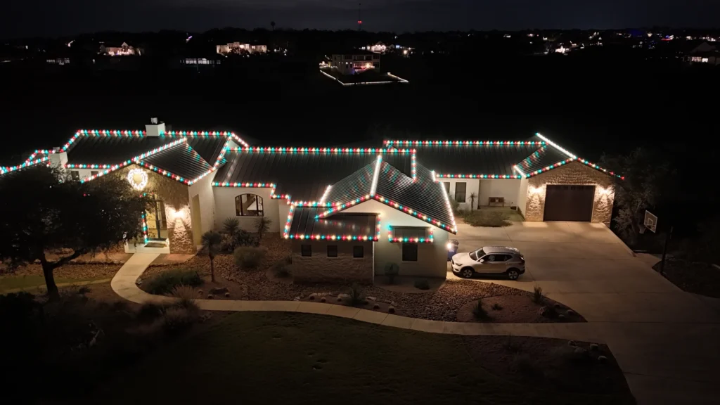 A large house is decorated with white, red, and green Christmas lights along the roof at night. A car is parked in the driveway, and other houses are visible in the dark background.