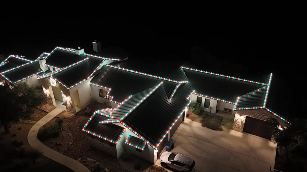A large house at night is outlined with festive red, white, and green holiday lights along the roof. A driveway with a parked car is visible, and the scene is well-lit against the dark sky.