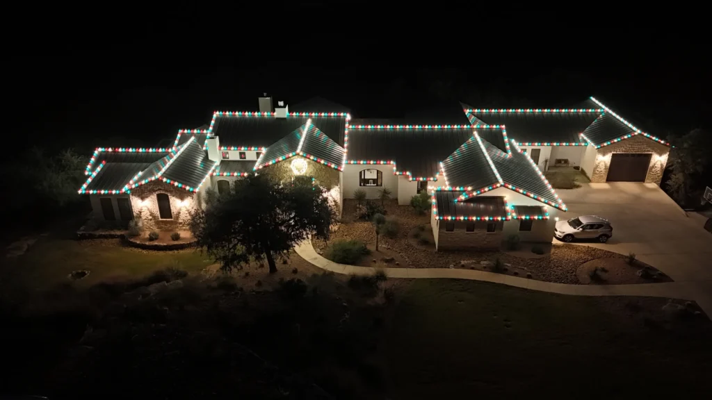 A large house at night, outlined with white and colored holiday lights along the roof, with a curved walkway and a tree in the front yard. The image is taken from above.