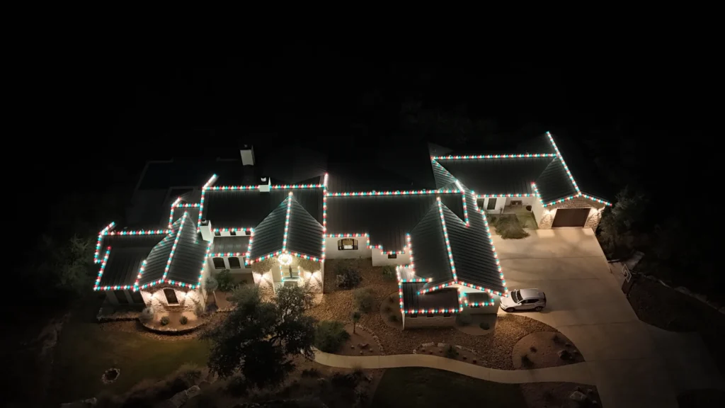 A large house is decorated with white and colorful Christmas lights along the rooflines, viewed from above at night. A driveway with a parked car and landscaped yard are visible.
