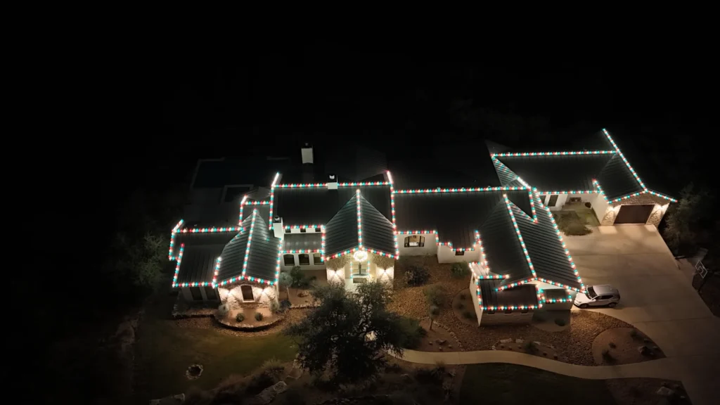 A large house is decorated with colorful string lights along the rooflines, glowing brightly at night. The surrounding yard and driveway are dimly lit, with trees and landscaping visible in the darkness.