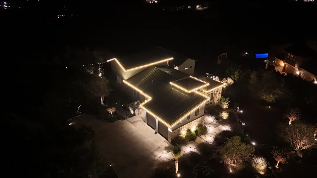 Aerial night view of a house with its roof outlined in warm white string lights, surrounded by trees and exterior landscape lighting, creating a festive and welcoming atmosphere.