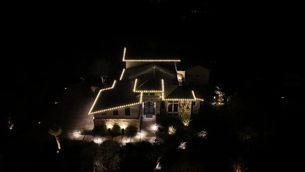 A house is outlined with warm white string lights along the roof, glowing against the night sky. The front yard and driveway are dimly lit, and nearby trees and bushes are partially visible in the darkness.