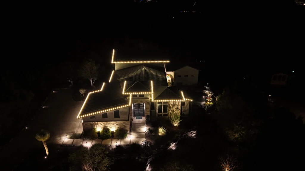 A house at night is outlined with bright white lights along the roof, surrounded by trees and landscape lighting that softly illuminates the yard and pathway.