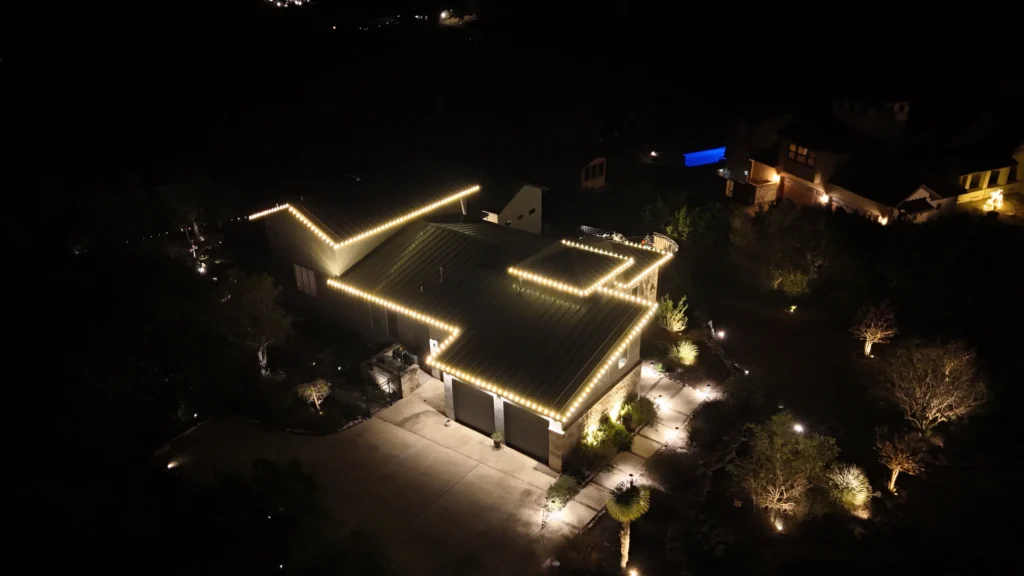 Aerial night view of a house outlined with warm white string lights along the roof. The driveway and surrounding trees are illuminated by outdoor lighting, with other houses faintly visible in the dark background.