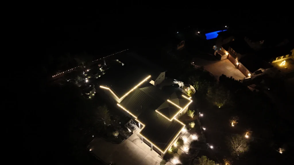 Aerial night view of a large house outlined with warm white lights, surrounded by trees and pathways illuminated by small lights, with a blue-lit pool visible in the background.