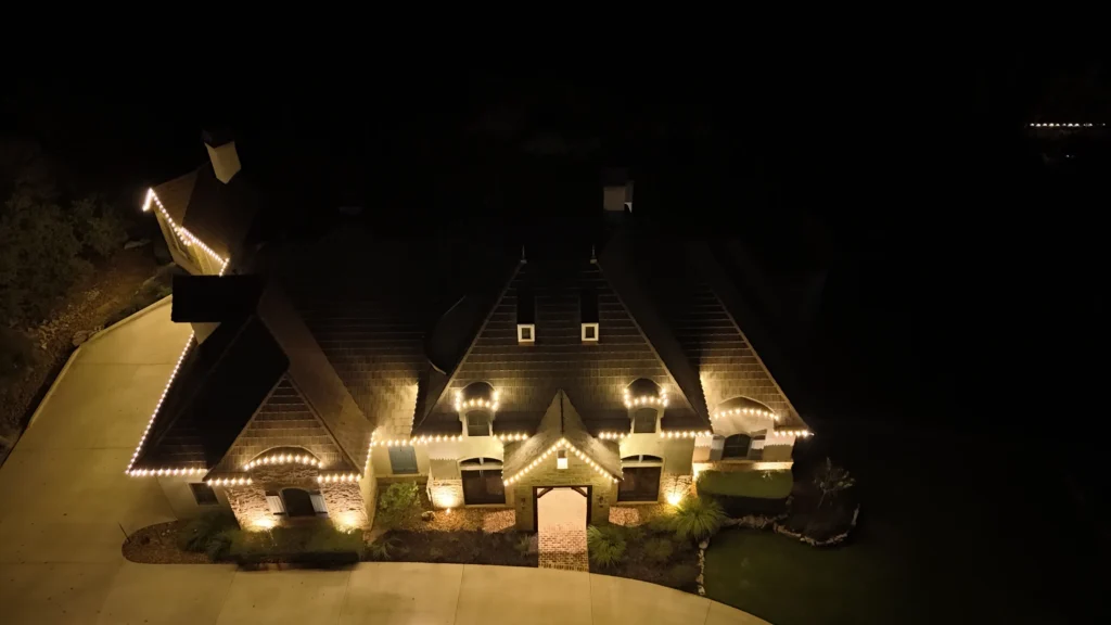 A large house is illuminated at night with warm white lights outlining the roof and windows. The property has a driveway, manicured lawn, and landscaping, all partially visible around the glowing home.