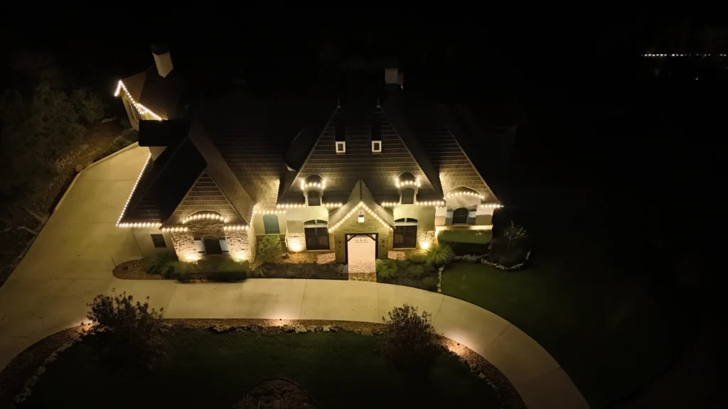 A large house is illuminated at night with white string lights outlining the roof, windows, and entryway. The curved driveway and landscaped yard are softly lit by ground lights. The surroundings are dark.