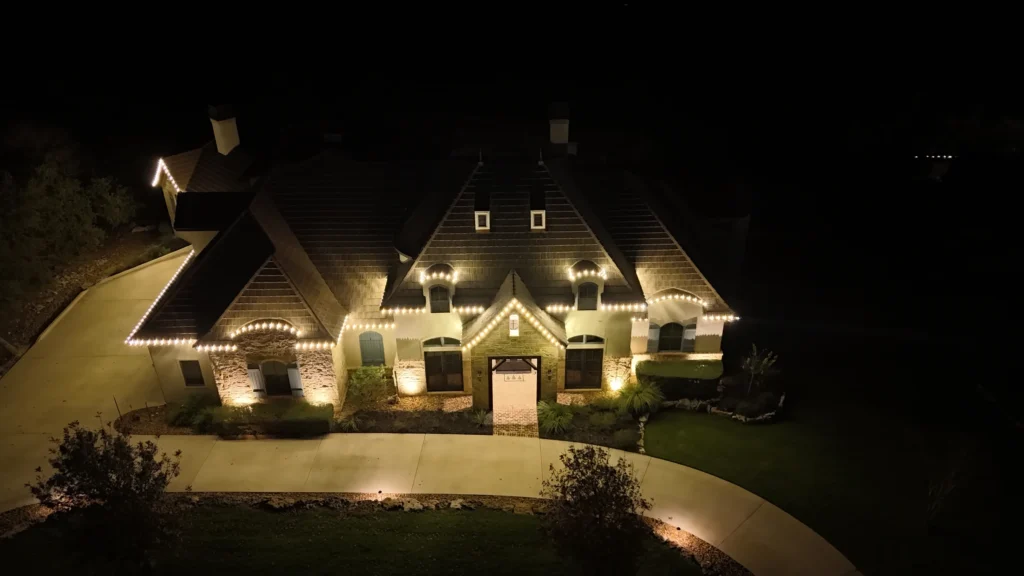 A large house is illuminated at night by white string lights outlining the roof and windows, with a curved driveway and neatly trimmed lawn in front. The surrounding area is dark.