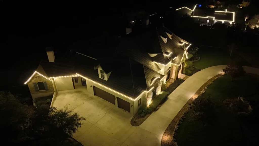 A large house at night with white holiday lights outlining the roof and garage, viewed from above. The driveway curves past the house, and another illuminated home is visible in the background.