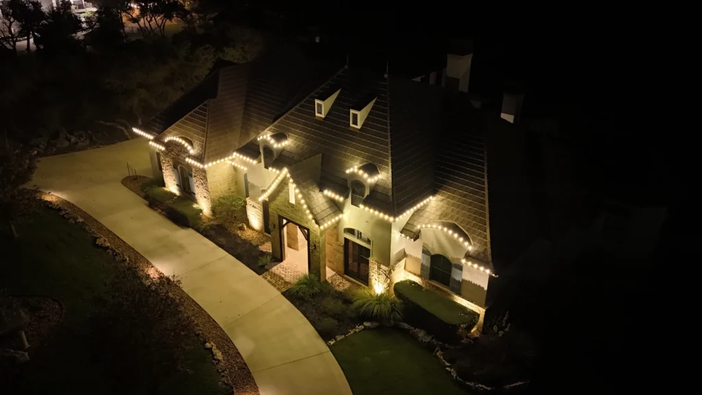 A large house with a steep, dark roof is decorated with white string lights along the roofline and front entrance. The house is surrounded by a curved driveway and a small lawn, illuminated at night.