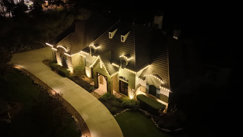 A house is outlined with warm white string lights along the roof and driveway at night, creating a festive and welcoming atmosphere. The surrounding yard and sidewalk are partially illuminated.