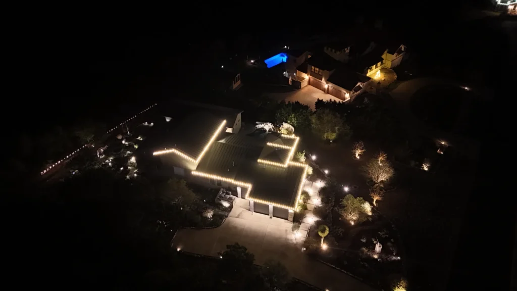Aerial nighttime view of a house with its roof edges outlined in white lights, surrounded by illuminated trees and pathways. Nearby, another home and a glowing blue-lit swimming pool are visible in the background.