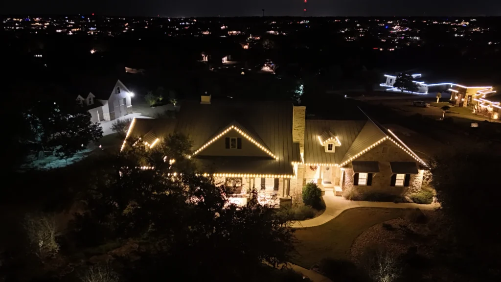 A large house decorated with white holiday lights along the roofline, photographed at night. The house is surrounded by trees, with neighboring homes and city lights visible in the background.