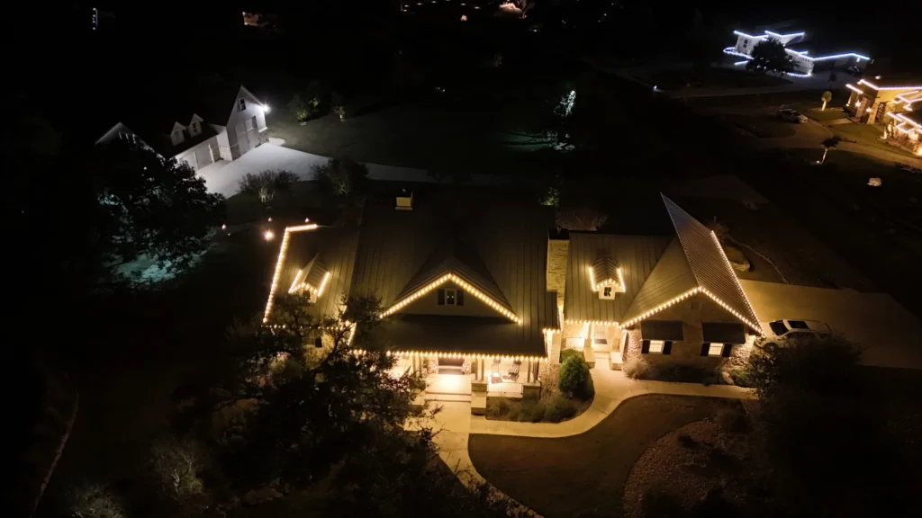 Aerial night view of a house with a dark roof outlined by warm yellow lights, surrounded by trees and winding walkways, with other illuminated buildings visible in the background.