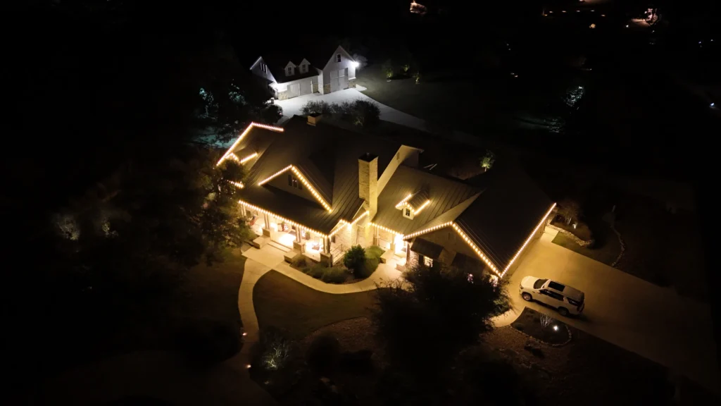 Aerial night view of a house decorated with warm white string lights along the roofline, with a car parked in the driveway and another illuminated house visible in the background.