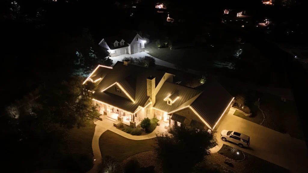 Aerial night view of a house with warm white lights outlining the roof and porch. A curved driveway leads to the entrance, and a car is parked nearby. Another illuminated house is visible in the background.