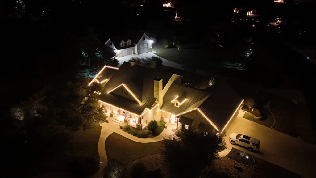 Aerial night view of a house with warm exterior lights outlining the roof, a curved driveway, and a parked car. Another illuminated house is visible in the background amidst trees.