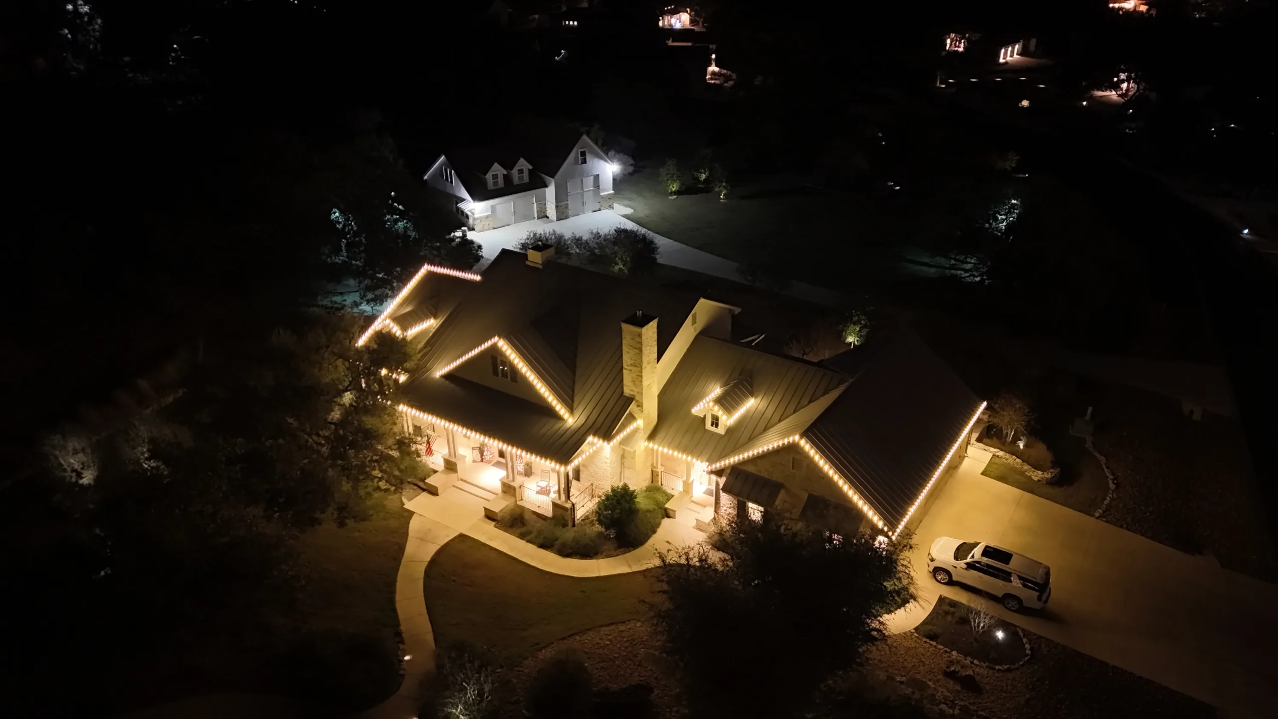 Aerial night view of a house with warm exterior lights outlining the roof, a curved driveway, and a parked car. Another illuminated house is visible in the background amidst trees.