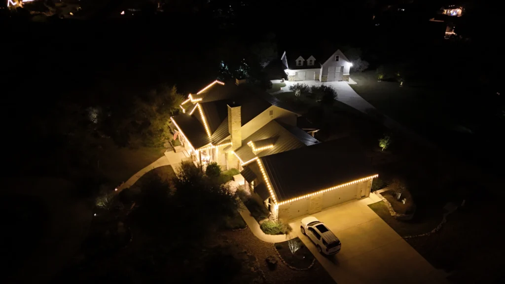 Aerial night view of a house decorated with white Christmas lights along the roofline and driveway. A white car is parked in front, and another building sits behind the house, both surrounded by trees and lawn.