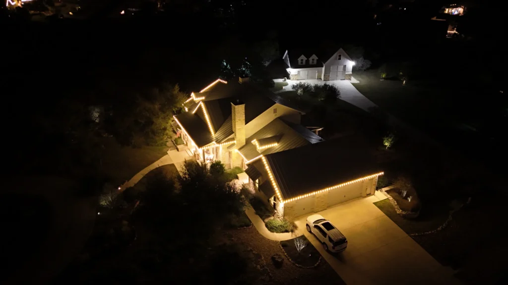 Aerial night view of a house decorated with warm white string lights along the roofline, a car parked in the driveway, and another illuminated building in the background.
