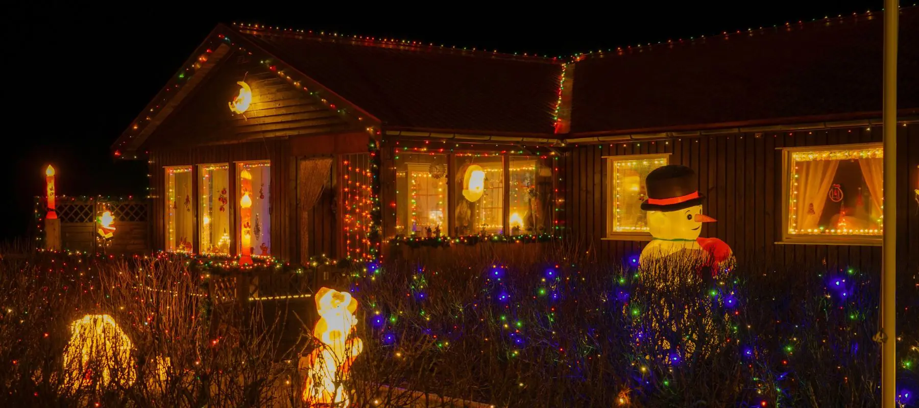 A house decorated with colorful Christmas lights, glowing candles in the windows, and an inflatable snowman in the yard at night. Shrubs in front are also adorned with festive lights.