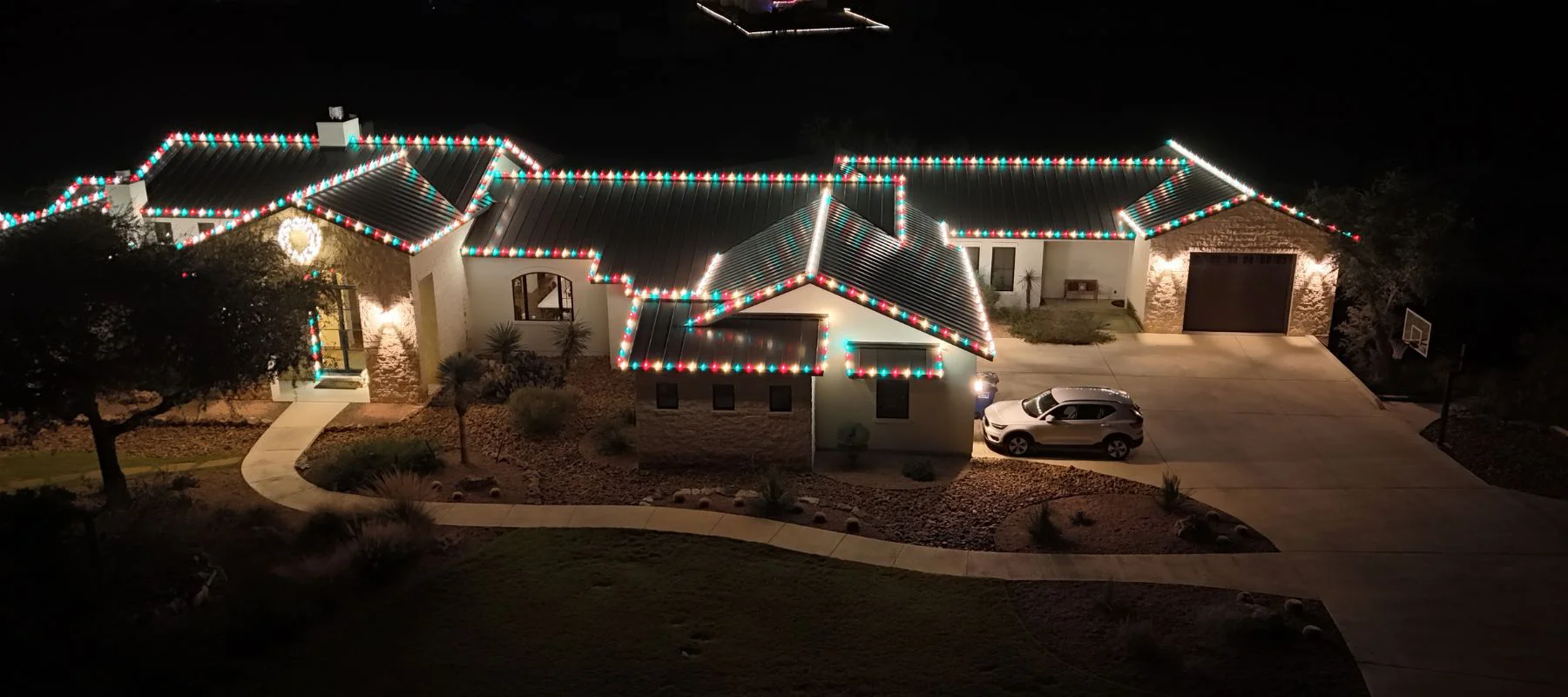 A large house at night is decorated with multicolored Christmas lights along the roof. A white car is parked in the driveway, and the front yard is landscaped with rocks and plants.
