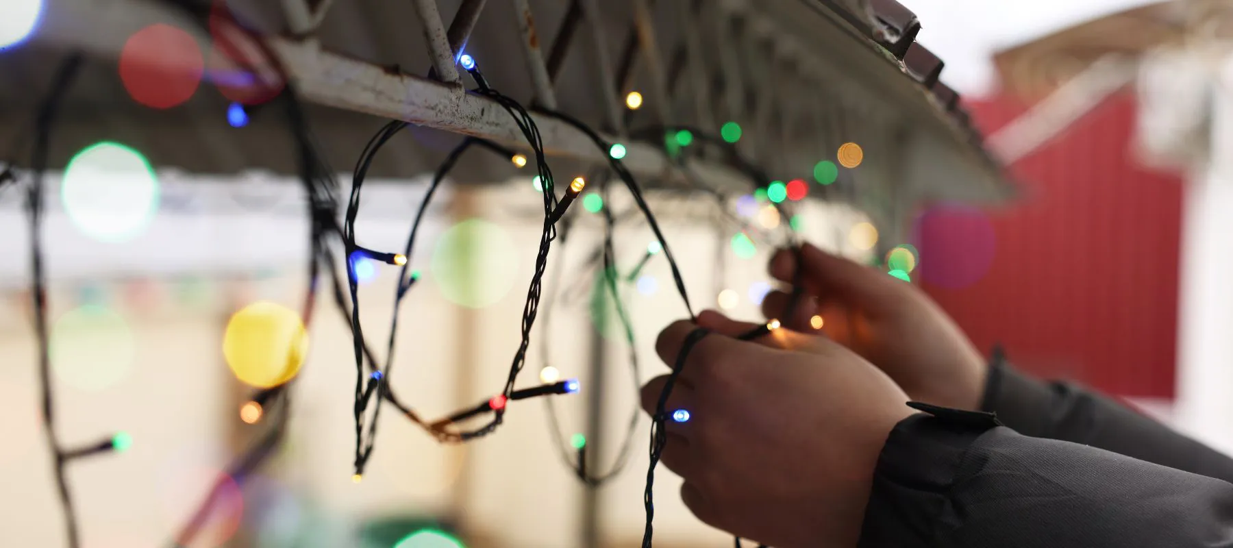 Close-up of a person’s hands hanging colorful string lights along the edge of a roof, preparing for decoration. The background is blurred with more lights visible.