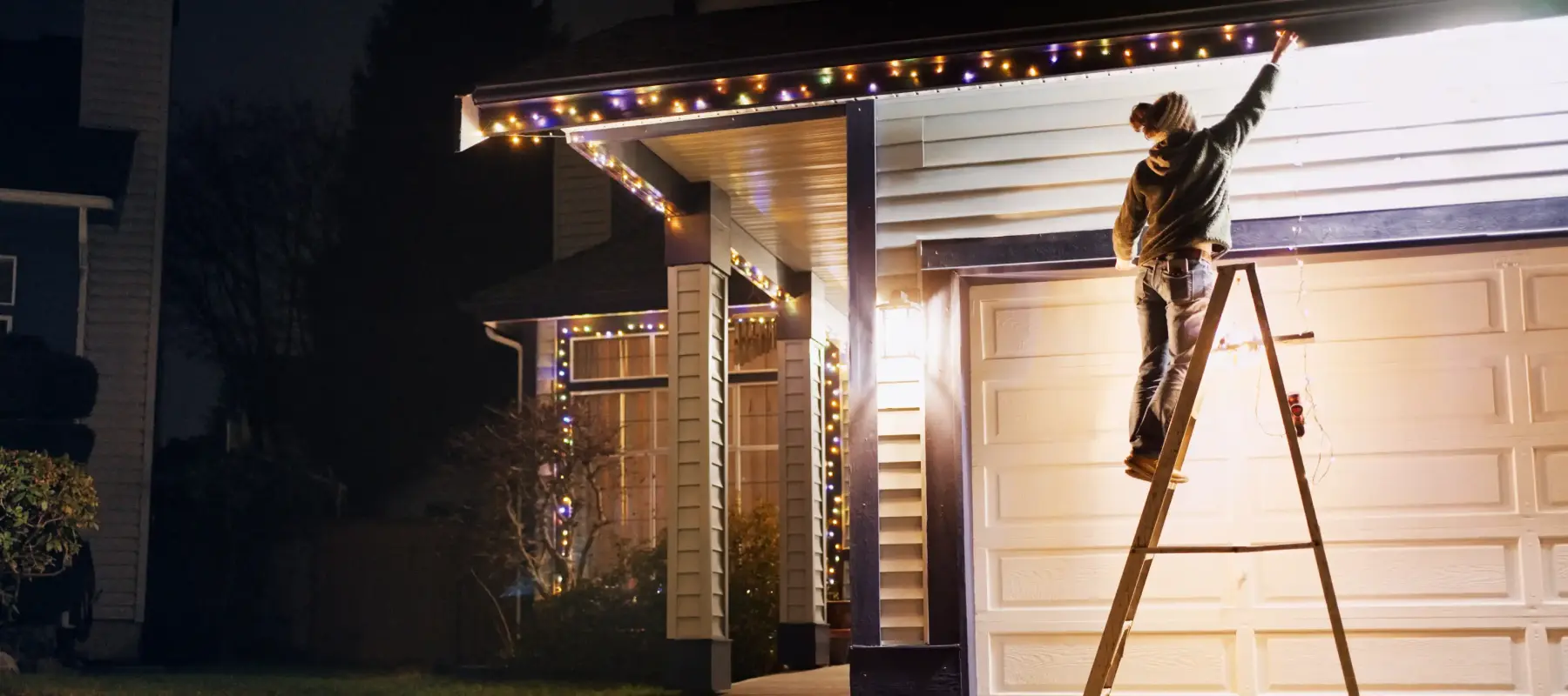 A person stands on a ladder hanging multicolored string lights along the edge of a garage roof at night, decorating the house for the holidays.